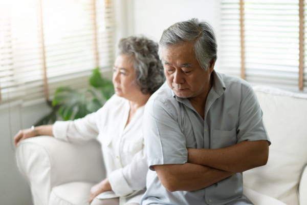 Upset couple with gray hair sitting on a white couch, woman looking straight ahead, man with his back to her and arms tightly crossed; concept is apology